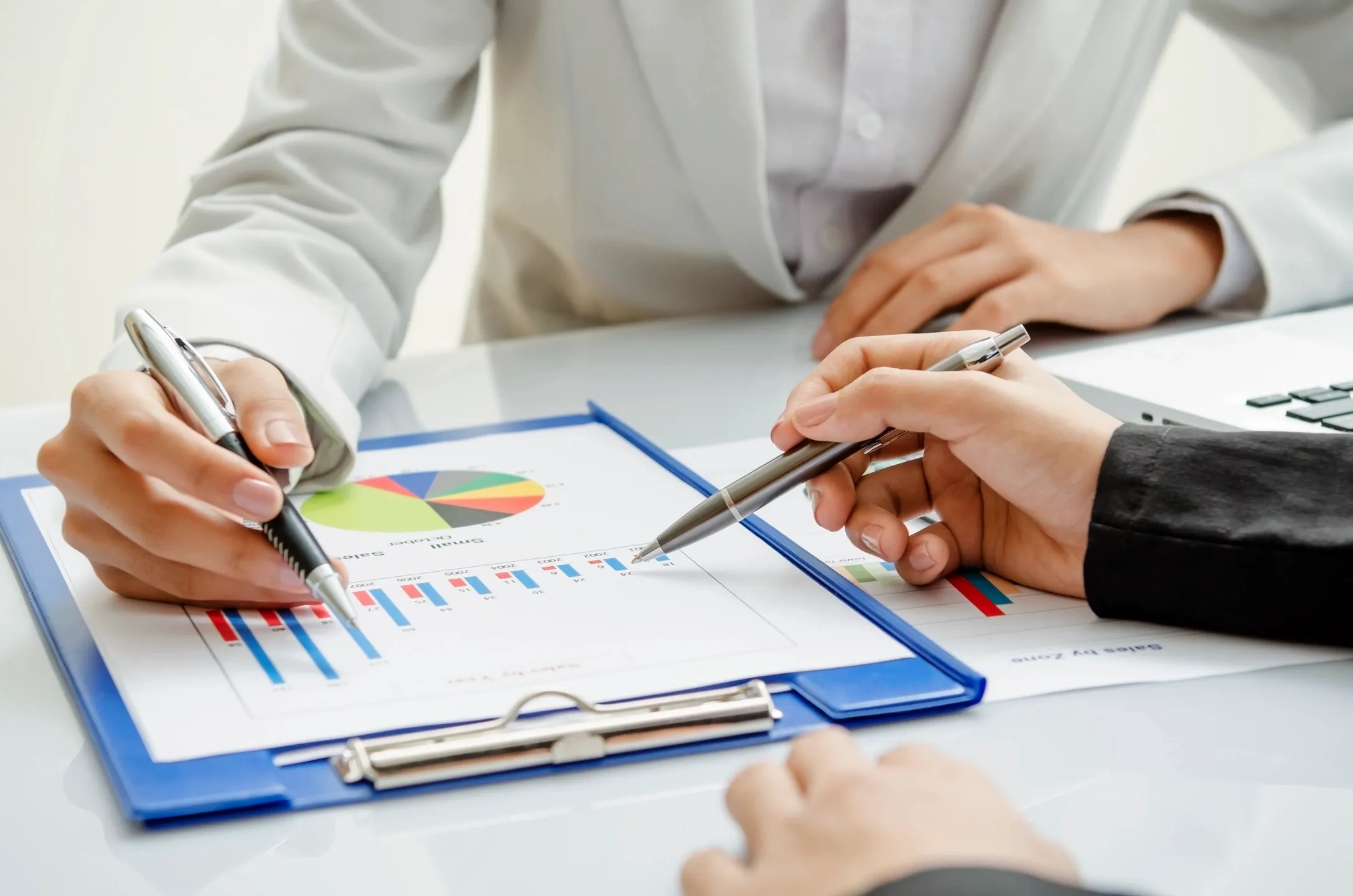 286 Two people in business attire sit at a table reviewing graphs and charts on a clipboard, discussing data with pens in hand.