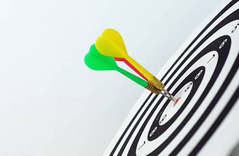 Close-up of two darts, one green and one yellow, hitting the bullseye of a black and white dartboard.