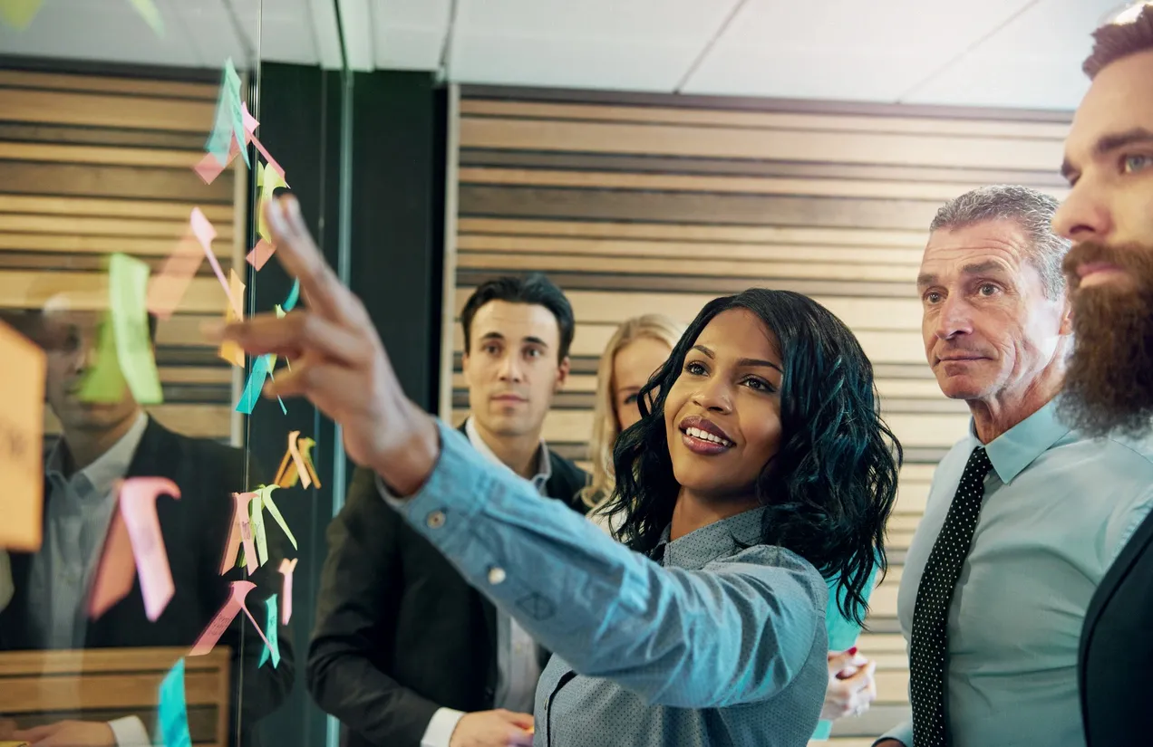 Q3VZ7AA A group of professionals in business attire stand by a glass wall with sticky notes, as one woman gestures and smiles while discussing ideas.