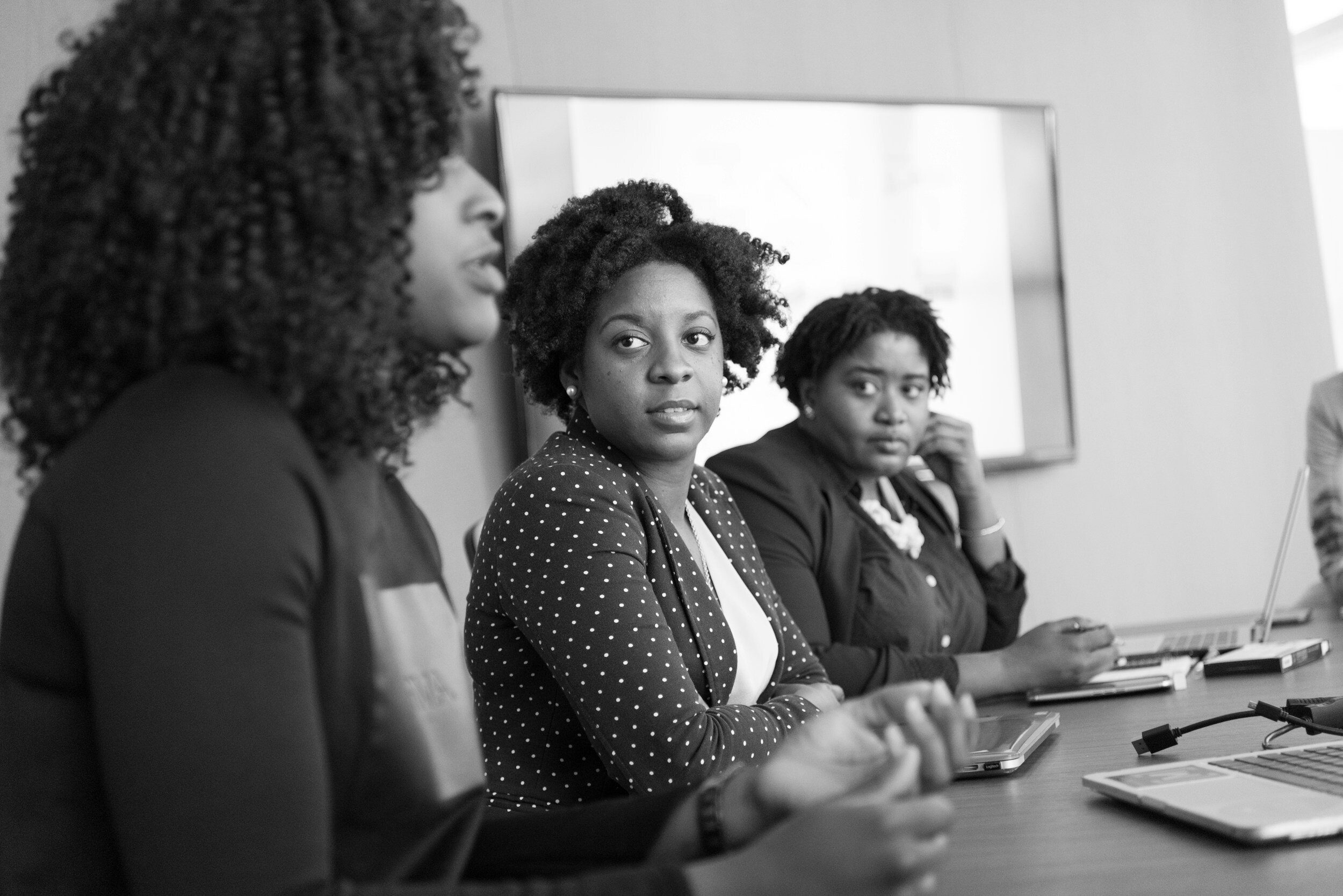 image-asset (1) Three women sit at a conference table in discussion, with laptops and papers in front of them in a meeting room.