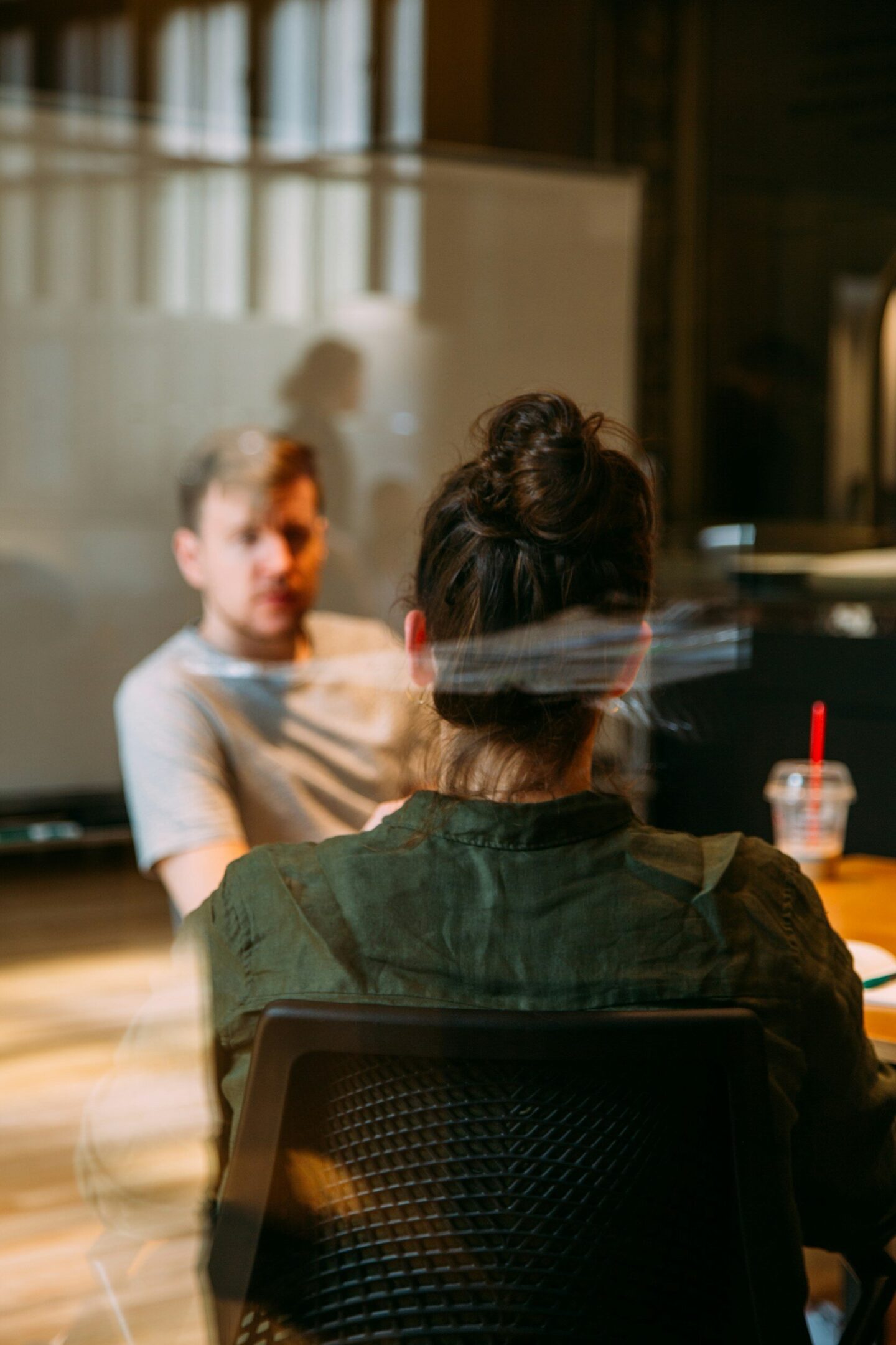 image-asset (2) Two people sit across from each other at a table in an office setting, one facing the camera with their back turned, and the other slightly out of focus.